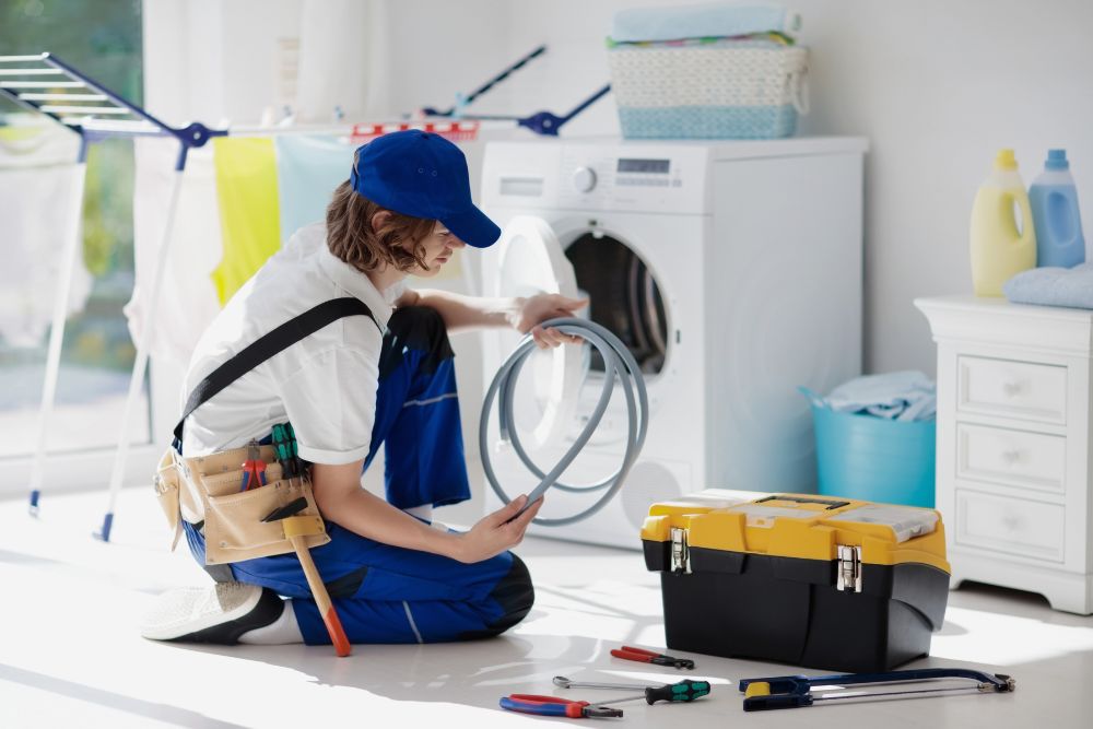 A woman in blue overalls is seated on the floor with tools, preparing to provide laundry appliance repair services in Honolulu, HI.