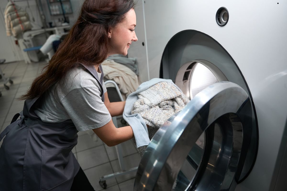 A worker in an apron loads towels into a large commercial washing machine in a clean, well-lit laundry facility.