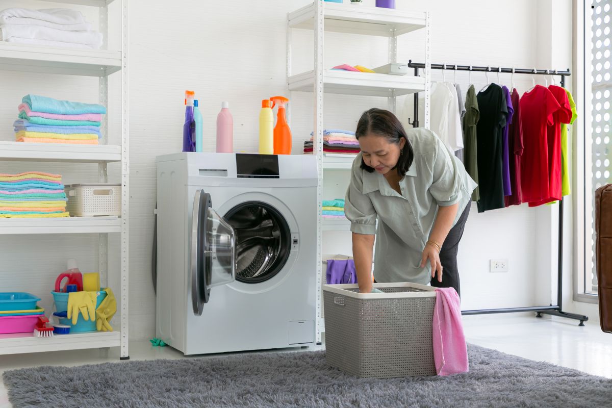 A person bends over a laundry basket in a bright laundry room, surrounded by colorful towels, cleaning supplies, and neatly arranged clothes.