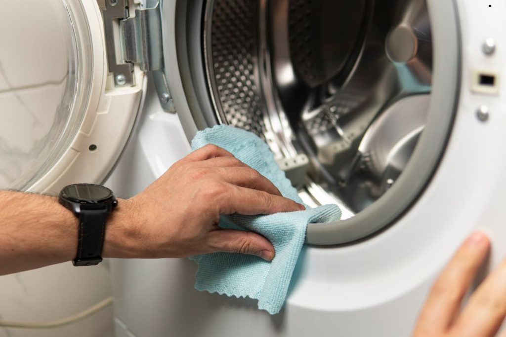 A person's hand with a black watch wipes the rim of a washing machine door with a light blue cloth.